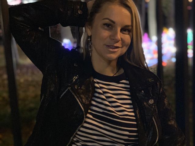 BelleLisaG con capelli biondi, che indossa una giacca di pelle nera e una camicia a righe, sorridente, con una mano dietro la testa, di notte, luci bokeh, piccoli orecchini, leggermente sorridente, casual, sexy..