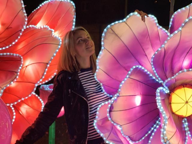 BelleLisaG, con i capelli biondi, indossa una giacca nera e una camicia a righe, ed è in piedi tra lanterne a forma di fiore, luminose e colorate, con bordi blu a LED, mentre guarda verso l'alto in un ambiente notturno..