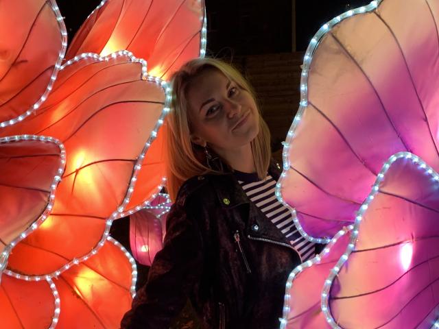 BelleLisaG, con i capelli biondi, indossa una giacca di pelle nera e una camicia a righe, ed è in piedi di fronte a luci a forma di farfalla, rosse e rosa, con bordi bianchi a LED..