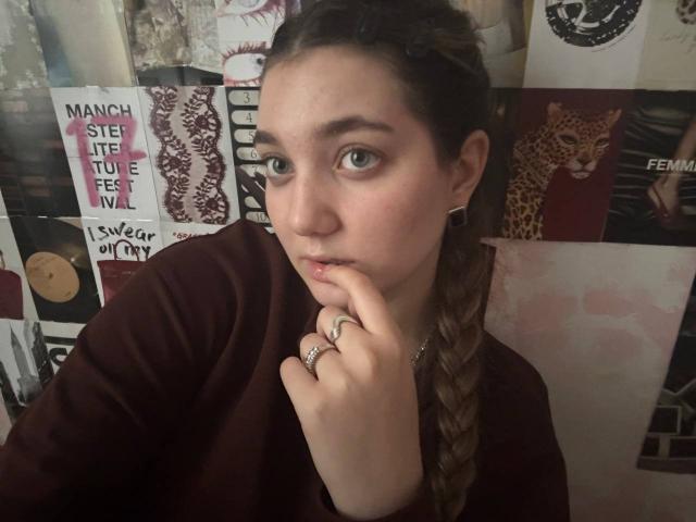 MichikoVivona with braided brown hair, green eyes, and a serious expression, wearing a maroon top, touching her chin with her finger, showing a ring on her finger.