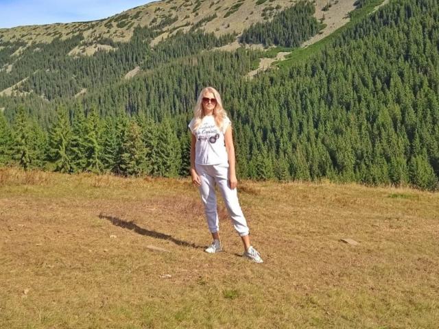 GladisBelle with long blonde hair, wearing white outfit, sunglasses, and blue sneakers, standing on grassy hill, forested mountain in background, sun shining, shadow on ground.