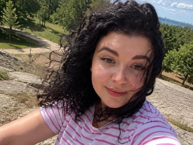 Yulia Rewiss hat lockiges, schwarzes Haar, einen Nasenring und trägt ein pinkgestreiftes Hemd. Sie lächelt mit dezentem Make-up und einem leichten Teint, in einer sonnigen Umgebung im Freien..
