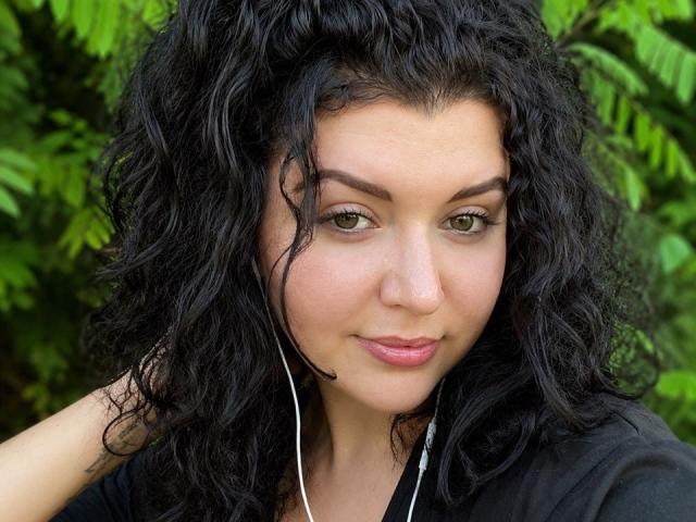 Yulia Rewiss, met krullend zwart haar, groene ogen en roze lippen, draagt een zwarte blouse, heeft witte oordopjes in en heeft een subtiele glimlach. Ze is buiten, omringd door groene begroeiing..