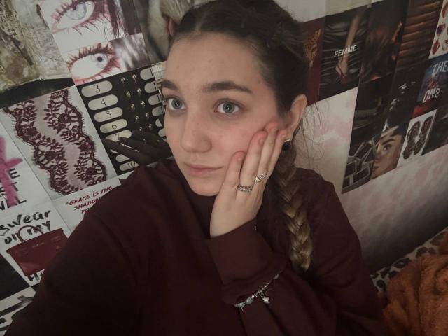 MichikoVivona has green eyes, a braid, and a hand touching her cheek. She's wearing a maroon top, with a ring on her finger, and a serious expression.