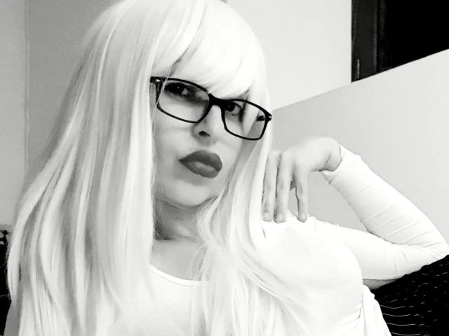 NattyNat-hot a de longs cheveux blancs et lisses, porte des lunettes noires et est vêtue d'un haut blanc à manches longues. Elle a une main posée sur son visage et regarde l'appareil photo..