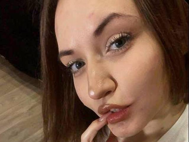 LelaTraverso has light brown skin, brown eyes, and is biting her finger with a slightly pouty expression. She has red-brown hair and is wearing a white shirt.