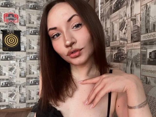 LelaTraverso with pale skin, brown hair, and pink lips, wearing a black top, showing her small tits and a silver bracelet, looking at the camera.