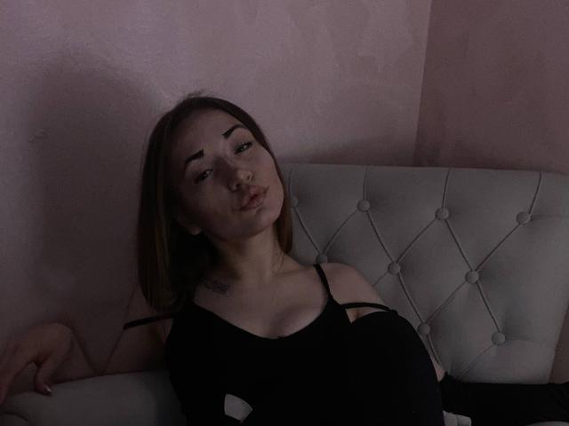 LelaTraverso with brown hair, black spaghetti strap top, small tattoos on chest, sitting on white tufted couch, looking up with slightly pouty lips, dimly lit pink wall.