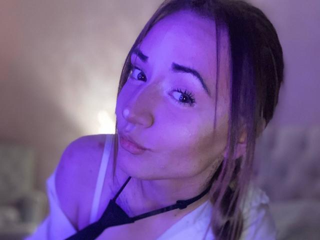 LelaTraverso with pouty lips, purple-lit face, and brown hair, wearing a black strap and white shirt, head slightly tilted to the side. Visible boobs and subtle makeup.