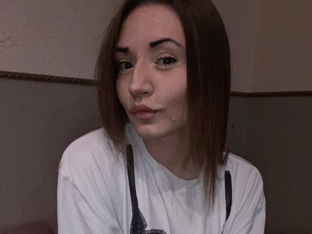 LelaTraverso with pouty lips, brown eyes, and straight brown hair, wearing a white shirt with black straps, in a dimly lit setting. Her face is slightly flushed.