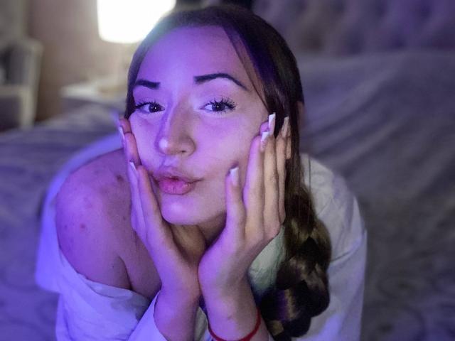 LelaTraverso with hands on cheeks, pouty lips, long brown hair in braid, white off-shoulder shirt, purple-blue lighting, visible freckles, and manicured nails, sitting on a bed.