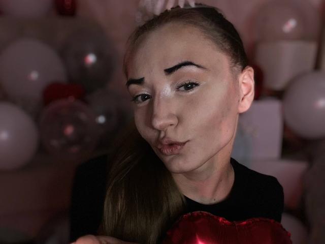 LelaTraverso with pouty lips, brown eyes, and eyebrows raised, wearing a black shirt with red heart, has her long brown hair in a ponytail, surrounded by balloons.