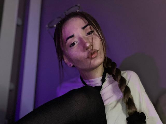 LelaTraverso with pouty lips, braided brown hair, glasses on head, white turtleneck, black sleeves, and black stockings, in dim purple light, looking slightly up.
