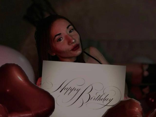 LelaTraverso with brown hair, dark lipstick, holding a birthday card, surrounded by pink heart balloons, looking at camera with a subtle smile.