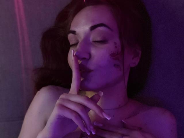 LelaTraverso with eyes closed, finger to lips, purple lighting, face marked with tattoos, long brown hair, nude, hand with manicured nails, subtle necklace, calm expression.