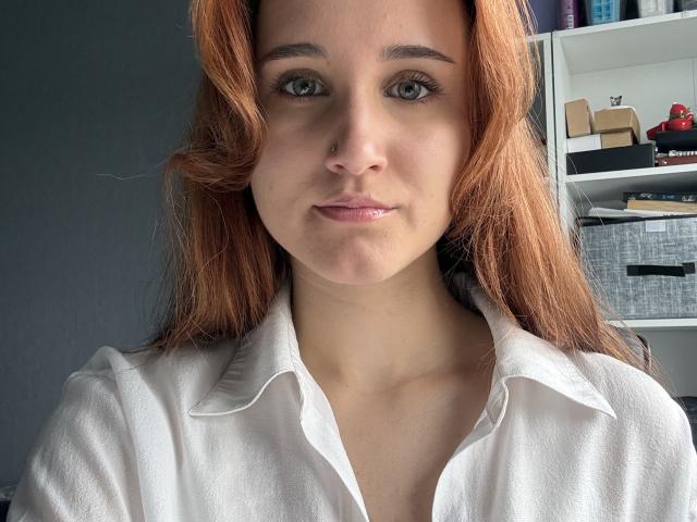 Gail Harmeson tiene el pelo naranja, ojos verdes, un piercing en la nariz y lleva una camisa blanca con un cuello ligeramente levantado. Sus labios rosados están ligeramente entreabiertos..