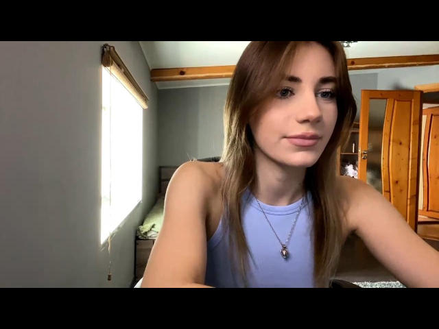 RosetteCerenzia with long brown hair, wearing a blue tank top, showing her tits, a small necklace, and a slight smile in a bright room.