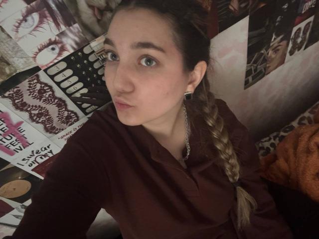 MichikoVivona with a long braid, wearing a maroon shirt, has blue eyes, small nose, and black ear gauges, looking into the camera, with a neutral expression.