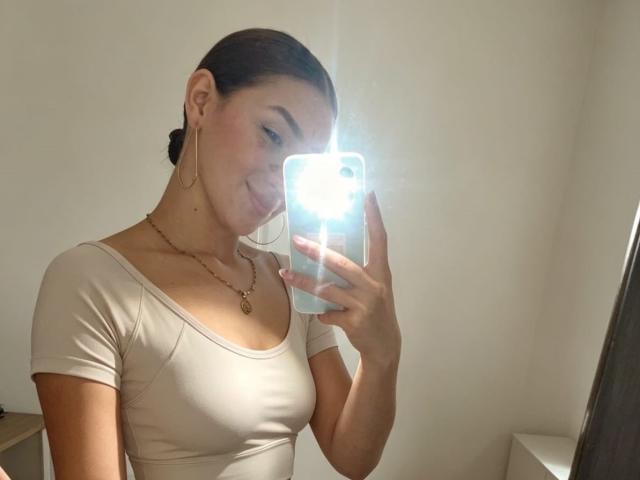 AbbyMilers with medium tits, smiling, holding phone, camera flash, beige top, gold necklace, earrings, dark hair in bun, light skin, mirror selfie, bright light.