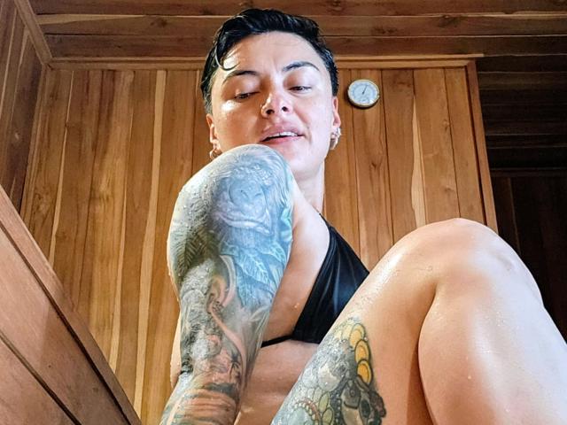 KatParadise mit kurzen, schwarzen Haaren, Tattoos an Armen und Beinen, schwarzem Bikini-Oberteil, Holzwänden, feuchter Haut, die nach unten schaut, lächelnd, mit Ohrringen, einer Uhr im Hintergrund, aufgenommen aus niedriger Perspektive..