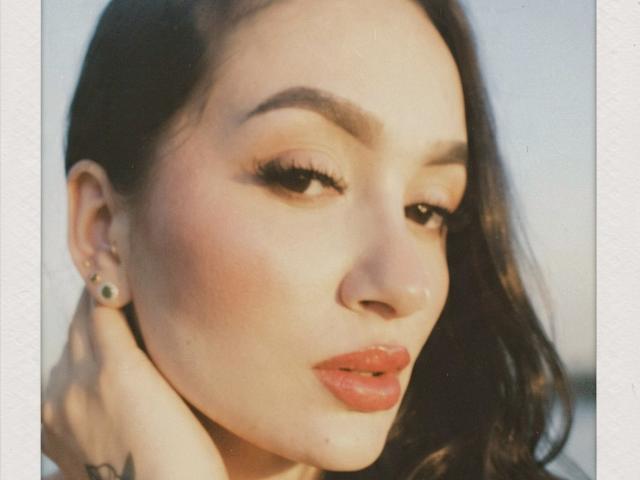 PamelaLiss a les lèvres pulpeuses, un rouge à lèvres rouge brillant, et elle touche son cou avec une main. Elle a les cheveux brun foncé, des boucles d'oreilles vertes, et elle regarde légèrement sur le côté..