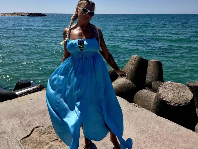 ChaudeEvely with blue dress, tits visible, standing by water, braided hair, white sunglasses, tattoo on arm, holding dress, sunlight, concrete, large black boulders, ocean in background.