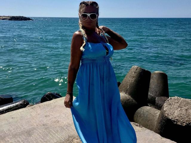 ChaudeEvely with dark skin, braided blonde hair, white sunglasses, blue sundress, holding dress, standing on concrete near ocean, rocks in background, clear blue water, sunny day.