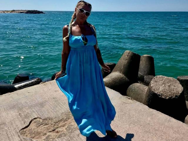 ChaudeEvely with blonde braids, wearing a blue dress, white sunglasses, standing on a rock by the ocean, holding her dress out, showing her tits. Ocean and rocks in background.