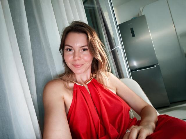Eva, con capelli castano di lunghezza media, sorridente, indossa un vestito rosso a fascia, è seduta e mostra le spalle scoperte e la parte superiore del petto, in una cucina con un frigorifero argentato..