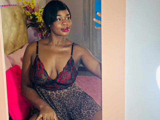 VenusWilly, avec une peau foncée, des cheveux noirs courts, du rouge à lèvres, portant une robe noire en dentelle et à motifs léopard, prenant un selfie devant un miroir avec un téléphone bleu. Seins visibles..