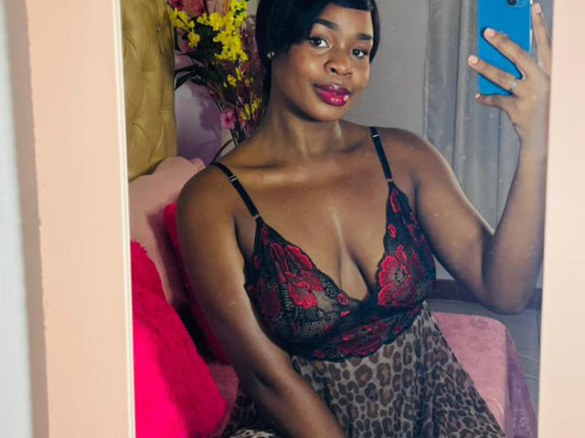 VenusWilly with dark skin, red lipstick, and black lace bra with red floral patterns, holding a blue phone, wearing a leopard print skirt, smiling in a mirror selfie.