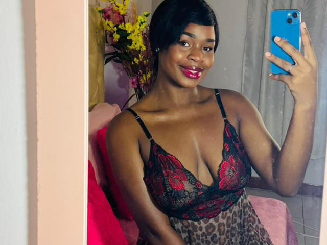 VenusWilly, avec sa peau foncée, son rouge à lèvres rouge et son soutien-gorge en dentelle noire, tenant un téléphone bleu, souriant, portant une robe à imprimé léopard, montrant ses grandes seins dans un selfie devant un miroir..