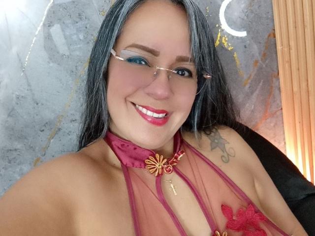 LailaCastillo with blue eyeshadow, pink lipstick, and glasses, smiling, wearing a sheer red halter top with a butterfly keyhole, exposing her tits, and a tattoo on her right shoulder.