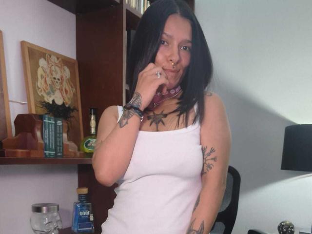 AmelieBlake with black hair, white tank top, nose ring, tattoos, pink choker, thinking with finger on lip, showing tits, seated, holding phone, shelf with liquor and art behind.