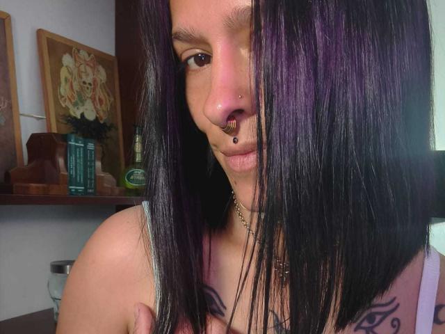 AmelieBlake with black straight hair, nose ring, and lip ring, showing off her tits with tattoos on her chest, holding her right boob, in a slightly dim room.