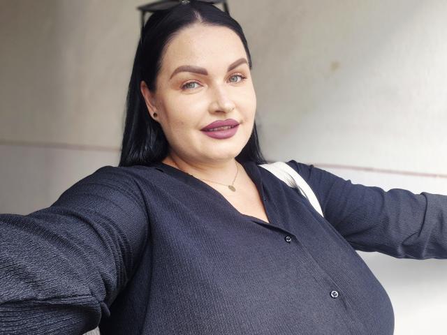 GeorgiaLerox has dark, straight hair, light skin, and is wearing a black, buttoned shirt. She has purple lipstick, a small necklace, and a confident, smiling face.