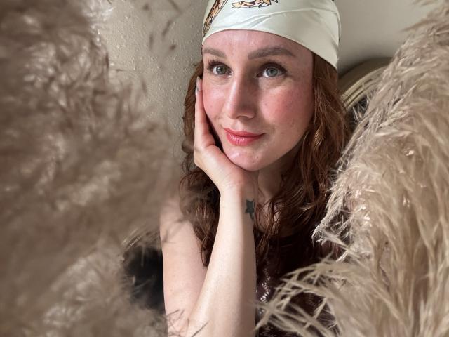 KatyaRogersX with red hair, white headband, pink cheeks, freckles, hand on cheek, looking at camera, soft brown furry texture in foreground, subtle smile, small tattoo on hand.