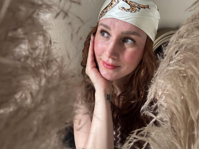 KatyaRogersX has red curly hair, a white headscarf with a gold pattern, fair skin, and a small tattoo on her right hand, gently touching her cheek. Fluffy beige fabric in foreground.