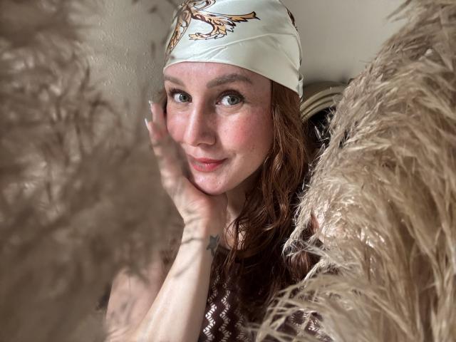 KatyaRogersX with red hair, white headscarf, and brown eyes, holding her face through fluffy beige curtains, showing a small tattoo on her hand. Soft smile, pink cheeks.