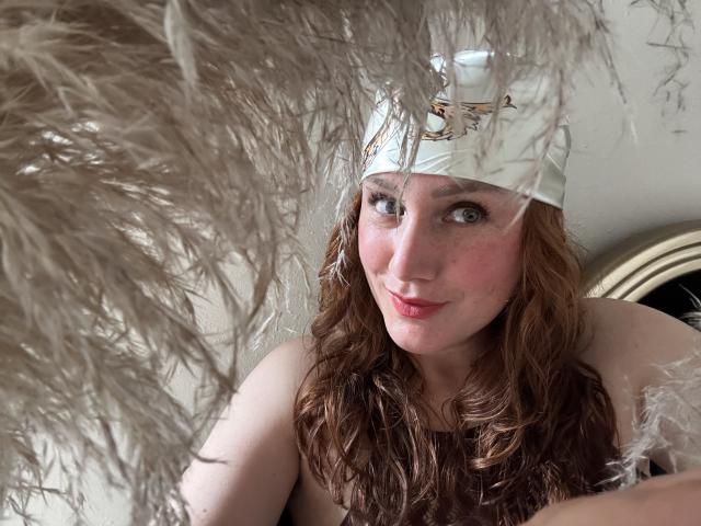 KatyaRogersX with wavy red hair, white feathered hat, and a cute smile, her tits partially covered by fluffy feathers in the foreground. Her pink lips and green eyes stand out.