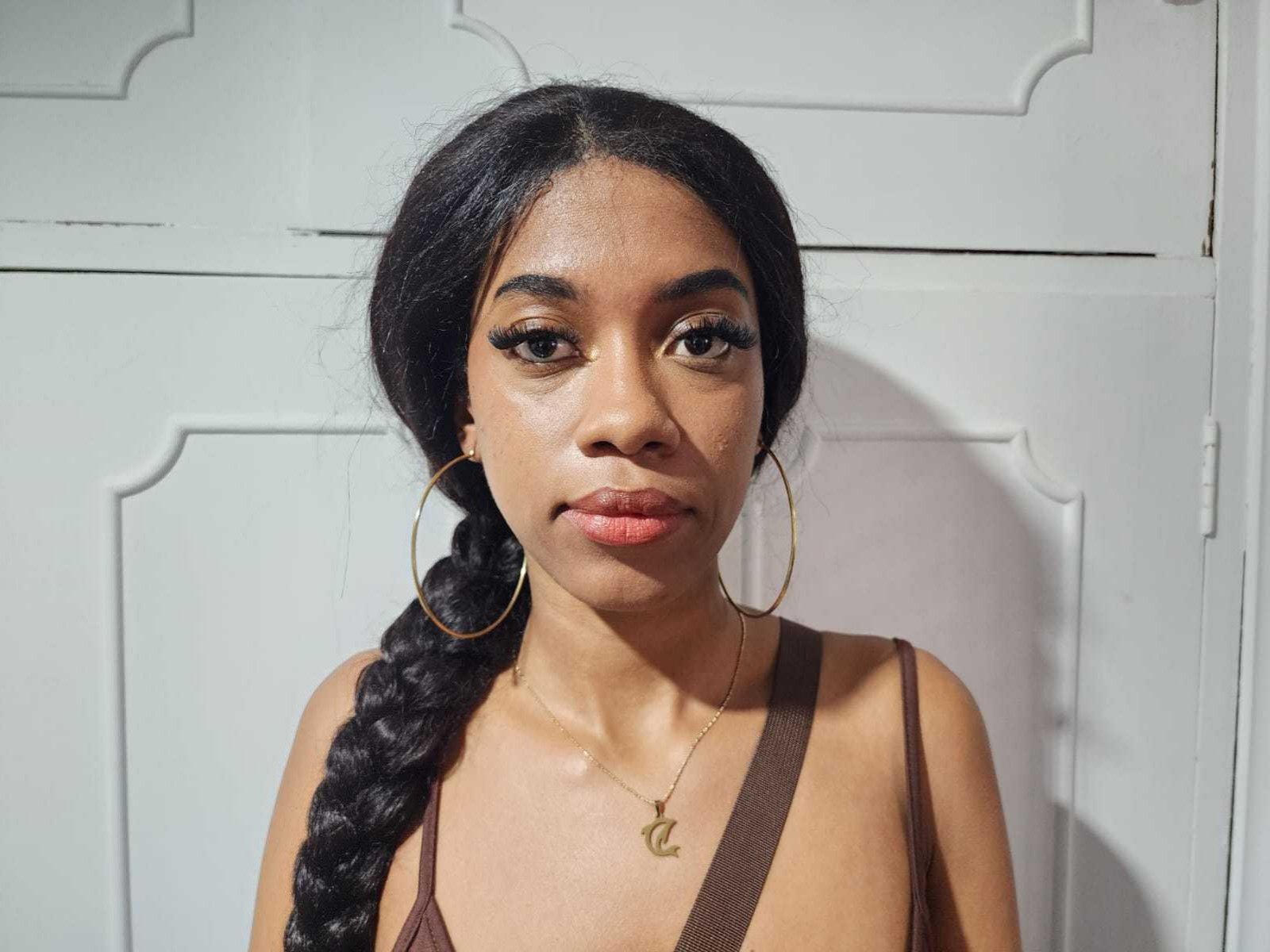 AllisonRomano a la peau foncée, une longue tresse noire, un débardeur marron, des boucles d'oreilles dorées et un collier en or avec un petit pendentif en forme de "Z". Elle a l'air sérieuse, avec les lèvres rouges légèrement entrouvertes..