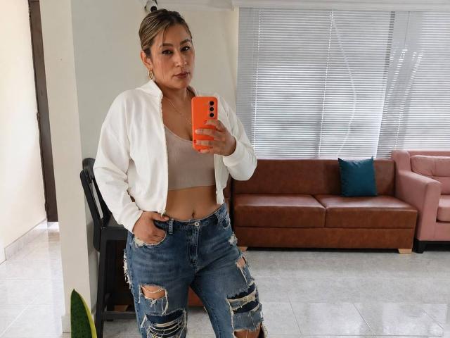 EmaFoxy, con la pelle abbronzata, i capelli castani raccolti in uno chignon, indossa una giacca bianca, un reggiseno beige e jeans blu strappati, tiene in mano un telefono arancione e si trova in una stanza, mostrando il suo sedere..