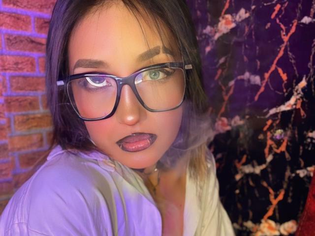 LunaixMoon has dark purple lipstick, black-rimmed glasses, and green eyes, with a white shirt, smoke in front, and dark hair, standing against a colorful, textured wall.