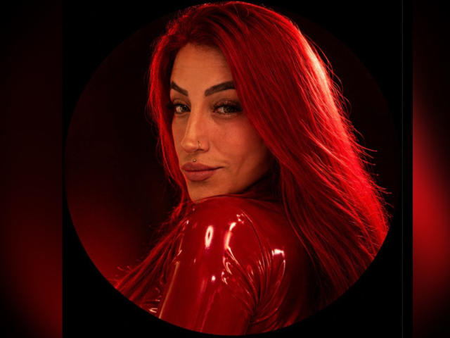 AlissaBrown with bright red hair, glossy red latex top, nose ring, and sultry look, turning to show her red tits in a circular frame against a dark background.