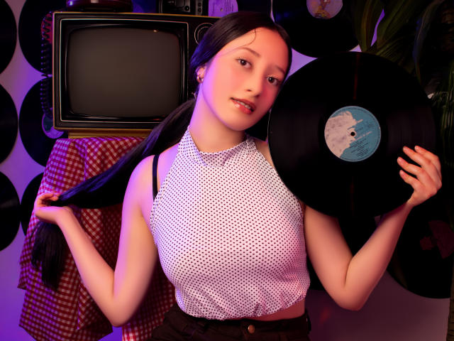 AmberBelle with long black pigtails, holding a vinyl record, wearing a white polka dot sleeveless top, standing in front of a retro TV, with pink and purple lighting.