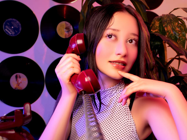 AmberBelle with red vintage phone, holding it to ear, wearing white polka dot sleeveless top, finger on chin, brown hair, pink-purple light, plant in background, records on wall.