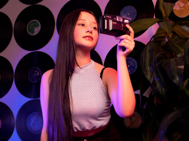 AmberBelle with long black hair, holding a camera, wearing a white polka-dot sleeveless top, purple-blue lighting, black vinyl records in the background, and a green plant on the right.