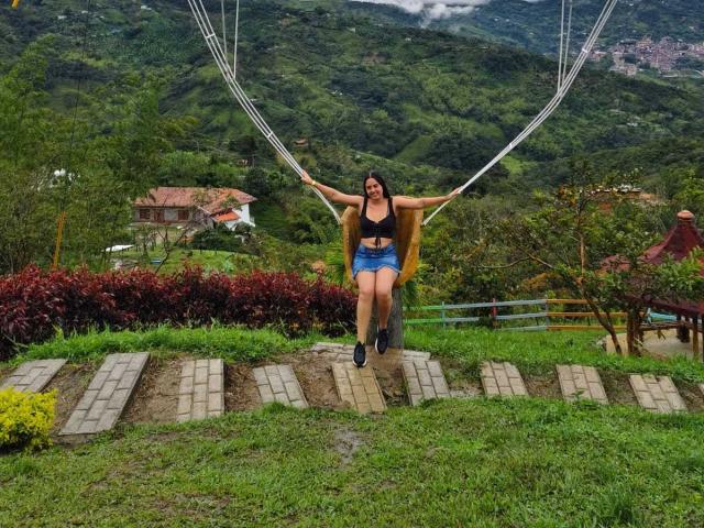 O traseiro da ZoeBlue está no ar, pendurada em um balanço de corda branca, vestindo uma cropped preta e shorts azuis, com colinas e árvores verdes ao fundo..