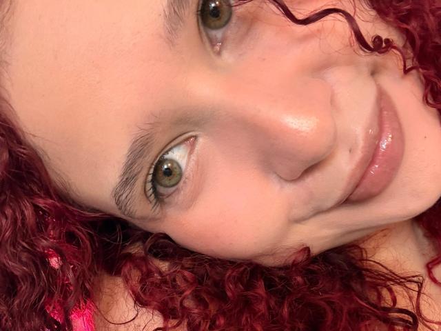 Il viso di ValeryScot, con i capelli rossi ricci, gli occhi verdi e un leggero sorriso. Le sue labbra rosa lucide e la pelle chiara sono visibili. Sta guardando verso l'alto con un'espressione carina e rilassata..