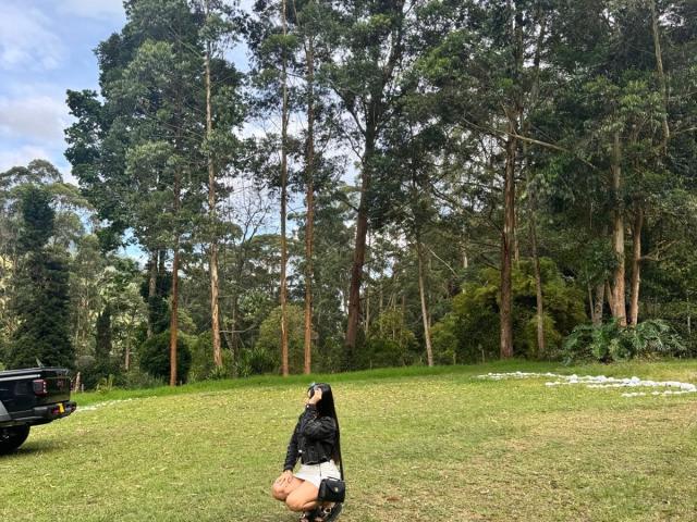 SophiaMarx kneels on grass, wearing a black jacket, white pants, and a hat, with a black truck on the left and tall trees in the background.